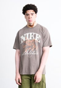 Grå bomulds t-shirt med et stort, falmet hvidt "NIKE" logo og en orange bevinget hest grafik, parret med grønne cargo bukser.