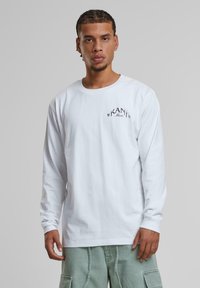 Weißes Langarm-T-Shirt mit kleinem schwarzem "KAN Music"-Logo auf der linken Brust, kombiniert mit hellgrünen Cargo-Hosen. Glatte Baumwolltextur.