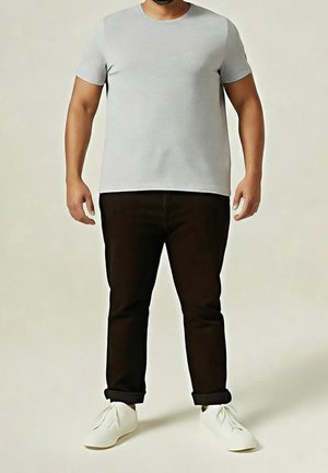 Man draagt een lichtgrijs T-shirt met korte mouwen, zwarte opgerolde broek en witte sneakers, staand tegen een neutrale achtergrond.