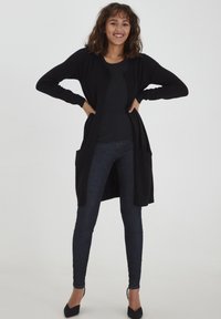 Zwarte lange cardigan met open voorkant, ribgebreide manchetten en twee voorzakken, gecombineerd met een zwarte top en donkere skinny jeans.