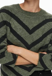 Pull en maille vert olive avec des rayures en chevrons noires. Il présente un col rond et des poignets côtelés. Texture douce avec une coupe décontractée.