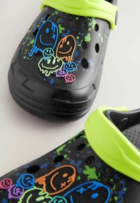 Schwarze Clogs mit einer strukturierten Oberfläche, die bunte Grafiken von Geistern und Smileys in Blau, Orange, Grün und Pink zeigen, mit einem hellgrünen Riemen.