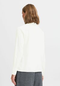 White long-sleeve blouse with a high neckline and button cuffs, made from a smooth fabric, shown from the back with a minimalistic design.