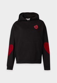 Sweat à capuche noir avec des patchs en forme de cœur rouges sur les coudes et un petit logo pomme rouge sur la poitrine avec le texte "DKNY" à l’intérieur.