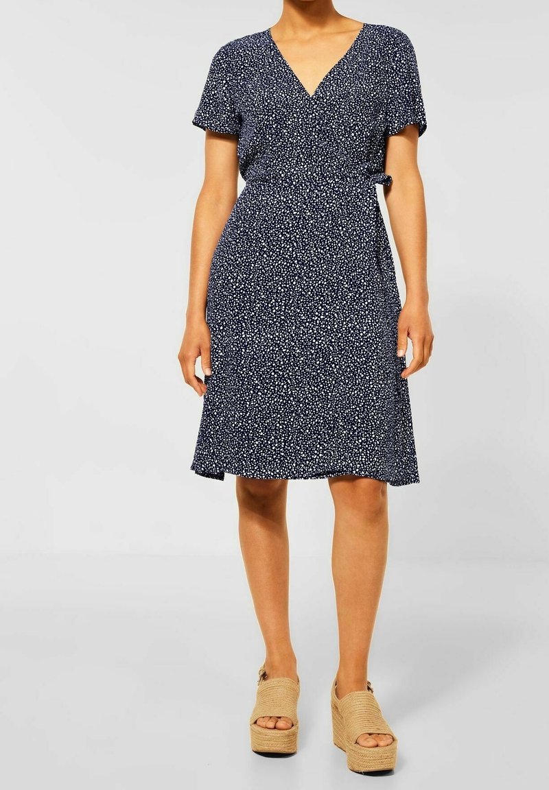 Femme portant une robe portefeuille bleu marine mi-longue avec des taches blanches et des sandales à plateforme beiges à bout ouvert, debout devant un fond uni.