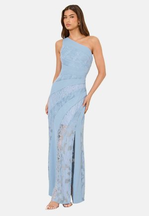 Frau trägt ein hellblaues, einseitig schulterfreies Spitzenkleid mit diagonalen Einsätzen und einem vorderen Schlitz, steht mit einer Hand in der Hüfte vor einem schlichten Hintergrund.