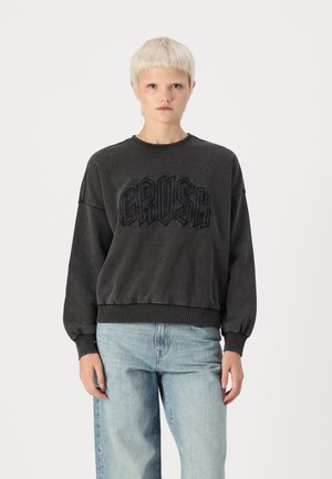 Gray sweatshirt with "CRUSH" in raised lettering, crew neck, ribbed cuffs and hem, paired with light blue jeans. Simple, casual design.