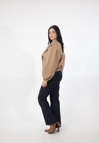 Braune kurze Jacke mit Schulterpolstern und elastischen Bündchen, kombiniert mit dunklen blauen Schlagjeans und braunen Stiefeln mit Absatz vor weißem Hintergrund.