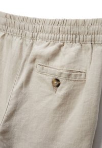 Shorts en lin beige avec une taille élastique et une poche arrière unique dotée d'un bouton rond et sombre. Texture lisse et coupe décontractée.