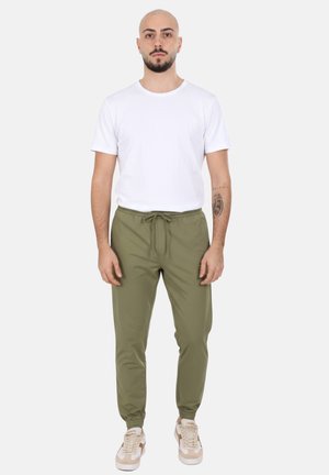 Uomo calvo con barba che indossa una maglietta bianca semplice, pantaloni da jogging verde oliva con coulisse e scarpe da ginnastica beige, in piedi davanti a uno sfondo bianco.