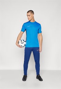 Blaues Sport-T-Shirt mit schwarzen Seitenakzenten, kombiniert mit blauen Jogginghosen mit einem orangefarbenen Nike-Logo und schwarzen Fußballschuhen, während ein Ball gehalten wird.