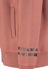 Rosa Sweatshirt mit weicher Textur, ausgestattet mit einer Seitentasche und bedrucktem Text entlang des Ärmels mit der Aussage: "Ich mag dich, weil du anders bist."