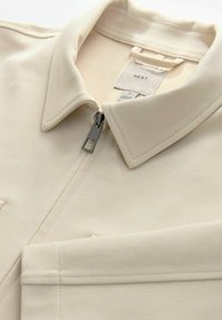 Kermansokkeluinen vetoketjullinen takki, joka on valmistettu teksturoidusta kankaasta, jossa on terävä kaulus ja hopeavetoketju. Tunnistuslabeli näkyy kauluksessa.