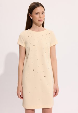 Kobieta z prostymi brązowymi włosami, ubrana w beżową sukienkę z krótkim rękawem ozdobioną małymi perłami i cyrkoniami na jednolitym jasnym tle.