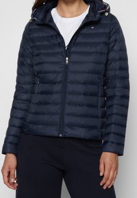 Kobieta w granatowej pikowanej kurtce z kapturem z zapięciem, pod którą ma na sobie białą koszulkę i ciemne spodnie.