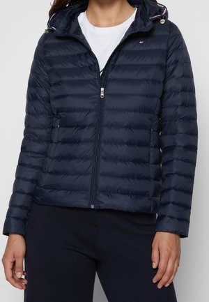 Kobieta w granatowej pikowanej kurtce z kapturem z zapięciem, pod którą ma na sobie białą koszulkę i ciemne spodnie.