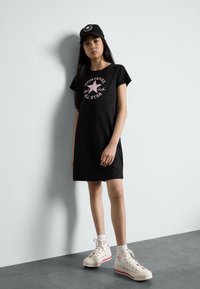 Sort kortærmet kjole med et pink Converse All Star grafik, parret med hvide high-top sneakers med røde detaljer. Enkel og afslappet design.