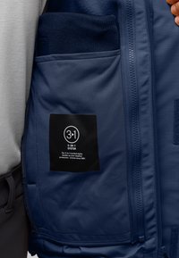 Interno della giacca navy con una texture liscia, con una tasca che contiene un tessuto navy spesso. L'etichetta descrive un sistema di protezione contro le intemperie 3-in-1.
