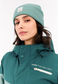 Türkiser Beanie mit gerippter Textur und Logo-Tag. Das Modell trägt eine passende türkise Jacke mit Knöpfen und einer Reißverschlusstasche.