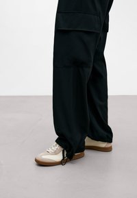 Personne portant un pantalon cargo noir avec des poignets élastiques et des baskets beiges avec des lacets blancs sur un sol gris.