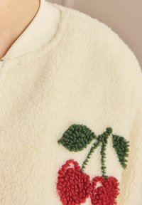 Cream fleece jacket with a zip, featuring red cherry embroidery and green leaves. Ribbed collar and soft, textured surface.