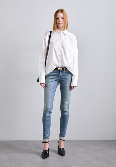 Cămașă albă cu guler și mâneci lungi, asortată cu blugi skinny de un bleu estompat și pantofi cu vârf ascuțit negri. Include un buzunar pe față și o geantă neagră.