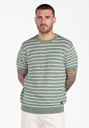 Man met kort haar en getatoeëerde armen draagt een groen-wit gestreept T-shirt met korte mouwen en een witte broek, staand tegen een witte achtergrond.