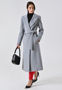 Cappotto grigio avvolgente con cintura auto-legante, colletto a scollo a V e maniche lunghe. Abbinato a leggings rossi e scarpe con tacco nere. Inclusa una borsa nera.