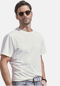 White short-sleeve t-shirt with a round neckline, paired with gray jeans and a textured belt; the model wears round sunglasses.