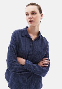 Blå rutig skjorta med knapp, krage, långa ärmar och en framficka. Tyget verkar mjukt med ett strukturerat mönster.