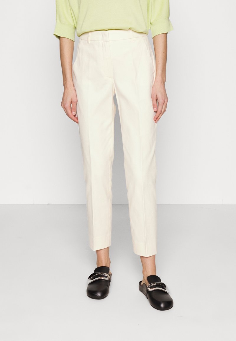Personne portant un haut vert clair, un pantalon beige clair ajusté aux chevilles et des chaussures noires à enfiler avec une boucle argentée.