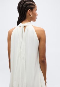 Kobieta z zaplecionymi warkoczami, zwrócona bokiem, ubrana w białą sukienkę bez rękawów, zawiązaną kokardą z tyłu na szyi, na jednolitym tle.