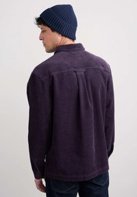 Violeta korduroja krekls ar aizmugures krokām, pogu aprocēm un brīvu piegriezumu, apvienots ar tumši zilu ribotu cepuri. Redzama teksturēta auduma detaļa.