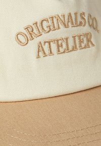 Crème cap met een tan kleurige klep, voorzien van geborduurde tekst "ORIGINALS CO. ATELIER" in lichtbruin. Gemaakt van gestructureerde stof.
