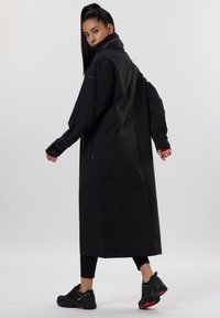 Černý dlouhý kabát s vysokým límcem, oversized rukávy a hladkou texturou. Nositelný přes černé legíny a spárovaný s černými botami.