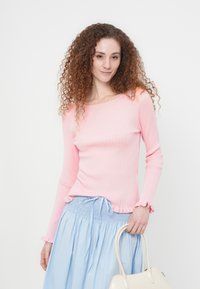 Νεαρή γυναίκα με σγουρά κόκκινα μαλλιά φοράει ένα ροζ ριπ μακρυμάνικο τοπ και ανοιχτό μπλε φούστα, κρατώντας μια λευκή τσάντα στο πλάι της.