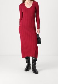 Robe rouge côtelée à manches longues avec un col rond et une silhouette droite, associée à des bottes noires montantes et un petit sac noir.