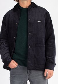 Mørkeblå kordfløjl jakke med knaplukning, krave og logo på venstre bryst. Båret over en mørkegrøn striktrøje, parret med grå jeans.