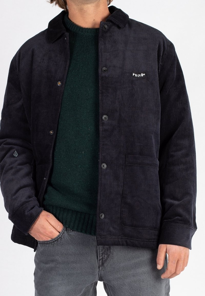 Mørkeblå kordfløjl jakke med knaplukning, krave og logo på venstre bryst. Båret over en mørkegrøn striktrøje, parret med grå jeans.