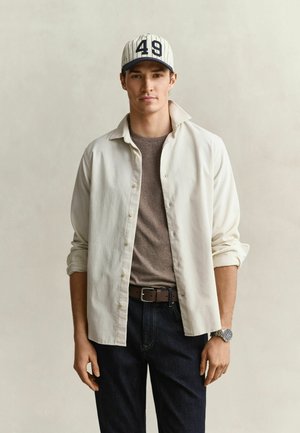 Jeune homme portant une chemise blanche boutonnée sur un t-shirt marron, un jean foncé, une casquette de baseball rayée avec le numéro 49 et une montre-bracelet.
