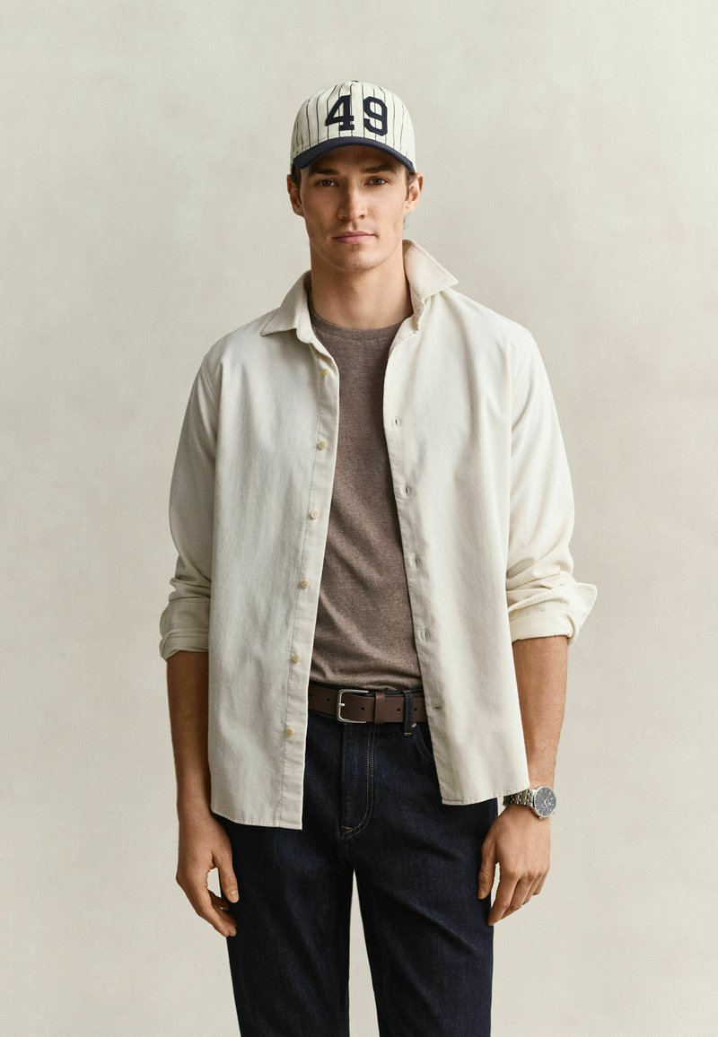 Young man wearing a white button-up shirt over a brown t-shirt, dark jeans, a striped baseball cap with number 49, and a wristwatch.
