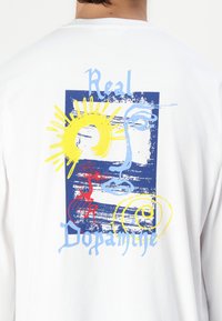 Cămașă albă cu mânecă lungă, cu un imprimeu grafic colorat ce reprezintă o față abstractă, soare și modele spiralate în nuanțe de albastru, galben și roșu.