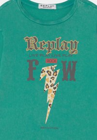 T-shirt de algodão verde apresenta um logo "Replay" cintilante, um design de raio em padrão de leopardo e texto em negrito. Textura suave, decote redondo.