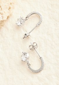 Pendientes de aro plateados que presentan una piedra preciosa clara y brillante montada con garras, con un diseño curvo y detalles texturizados a lo largo del aro.