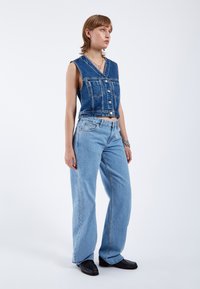 Chaleco de mezclilla con escote en V y botones en la parte delantera, combinado con pantalones vaqueros de pierna ancha de color azul claro. El conjunto presenta un ajuste casual y relajado, con una textura clásica de mezclilla.