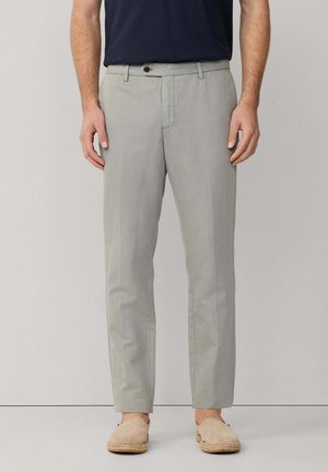 Hombre que lleva pantalones claros hechos a medida, camisa azul marino y alpargatas beige sin cordones, de pie frente a un fondo liso.