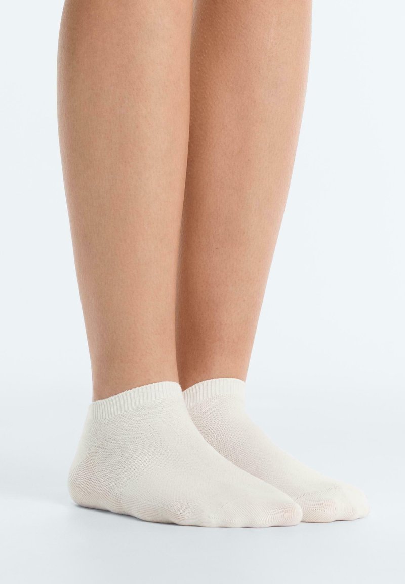 Jambes portant des chaussettes basses blanches aux chevilles, debout sur une surface blanche avec un fond clair.