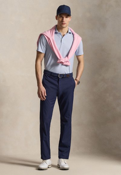 Νέος άνδρας που φοράει μπλε σκουφί, ανοιχτό μπλε πόλο πουκάμισο, ροζ πουλόβερ περασμένο στους ώμους, μπλε παντελόνι και λευκά παπούτσια, στέκεται μπροστά σε μπεζ φόντο.