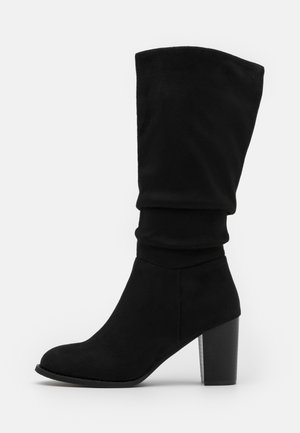 Botte noire pour femme mi-mollet avec talon bloc, bout arrondi et tige supérieure souple et flasque, vue de côté sur fond blanc.