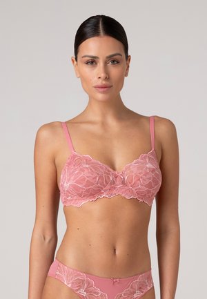 Donna con capelli scuri in reggiseno di pizzo rosa e biancheria intima coordinata, in piedi davanti a uno sfondo neutro e semplice, rivolta verso la fotocamera.